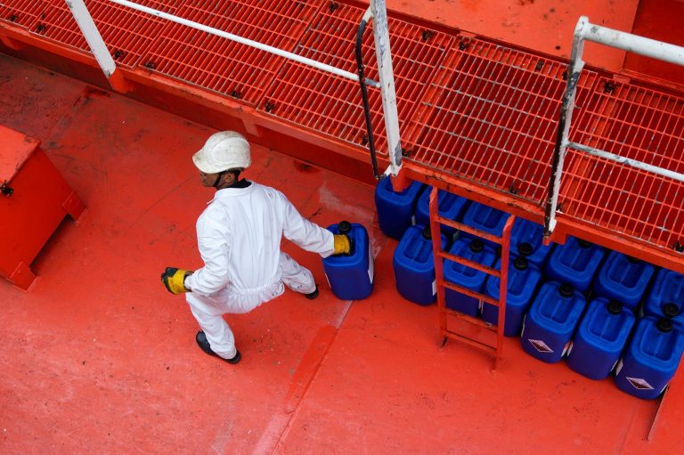 A maritime crew member wearing PPE moves blue fuel containers on a vibrant red ship deck.